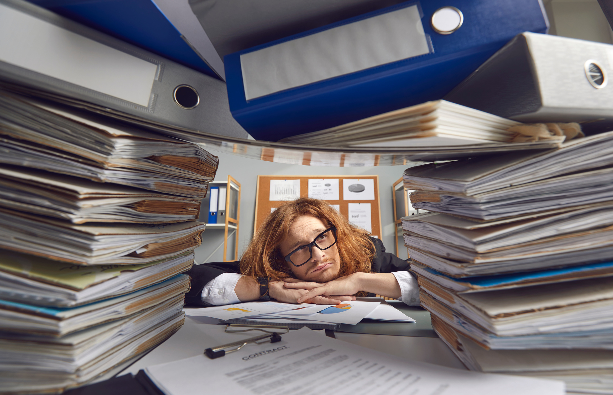 Tired Office Worker Sitting at Desk and Looking Apathetically at Piles of Paperwork